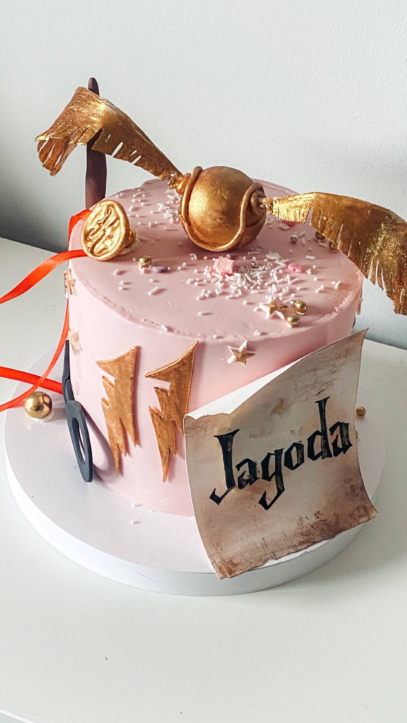 Różowy tort urodzinowy z dekoracją Złotego Znicza i napisem 'Jagoda' w stylu Harry'ego Pottera.