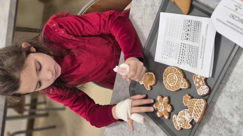 Dziecko w czerwonym ubraniu dekoruje pierniki lukrem podczas warsztatów cukierniczych.